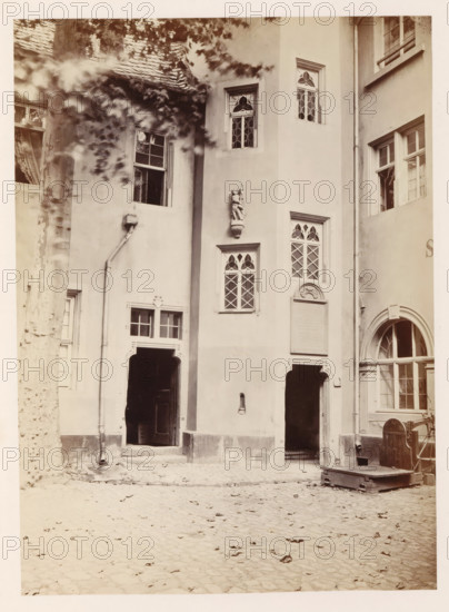 Exterior of the Zum Humbrecht house in Mainz, photograph, page, number: '18', photographer: B. Erdmann, Mainz, c. 1867 - in or before 1872, Rhineland-Palatinate, paper, albumen print, height 223 mm x width 164 mm, housing, façade (of house or building), A sepia-toned photograph of a courtyard façade showing several arched and rectangular doorways and windows set in smooth plaster walls, with decorative carved window frames and diamond-patterned leaded panes; a sculpted figure mounted on the wall above a small window; a drainpipe running down the wall beside a tall tree trunk with dappled light and blurred leaves in the foreground; cobblestone ground with scattered leaves; an open dark doorway leading into shadow, another arched entrance with panes of glass, and a wooden platform or lift with metal fittings positioned near the right side.