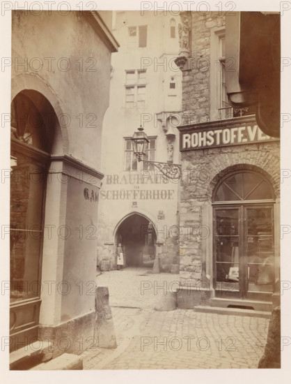 Exterior of Peter Schöffer's printing house in Mainz, photograph, page, number: '19', photographer: B. Erdmann, Mainz, c. 1867 - in or before 1872, B. Erdmann, photographer, Rhineland-Palatinate, paper, albumen print, height 235 mm x width 177 mm, printing, printing office, façade (of house or building), A narrow cobblestone alley flanked by textured stone and plaster buildings with arched doorways and windows, a decorative wrought iron street lamp projecting from a wall, a carved stone figure mounted on a corner, a glass storefront with wooden framing and visible interior shelves, a rounded arch passage leading further into the alley where a person in a light apron stands near the entrance, steps and low stone bollards at the walkway edges, and overhanging architectural elements casting soft shadows across the scene.