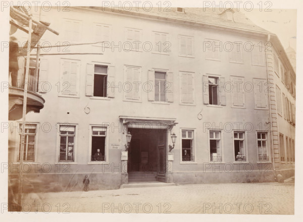 Front facade of the Gensfleisch family home in Mainz, photograph, page, number: '5', photographer: B. Erdmann, Mainz, c. 1867 - in or before 1872, Rhineland-Palatinate, paper, albumen print, height 173 mm x width 240 mm, housing, façade (of house or building), A multi-story building façade with rows of shuttered windows and a central open doorway flanked by decorative lamps, a small canopy above the entrance, and railings at the lower windows; several open windows reveal seated figures visible through the frames, a person standing near the left entrance window and another near the right; a curved balcony structure and vertical wooden poles are visible at the far left edge, and a cobblestone street runs along the front of the building.
