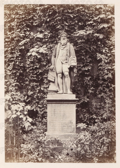Statue of Johannes Gutenberg in Mainz, photograph, page, number: '6', photographer: B. Erdmann, Mainz, c. 1867 - in or before 1872, Rhineland-Palatinate, paper, albumen print, height 197 mm x width 140 mm, sculpture, A stone statue of a bearded man stands on a rectangular plinth surrounded by dense foliage; the figure wears draped clothing with a long braided element over the shoulders and holds a folded scroll in one hand while a book rests near the other hand, the statue's legs are exposed below knee-length trousers and the base shows weathered surfaces, all set against a backdrop of climbing leaves and plants with foliage extending around and above the monument.