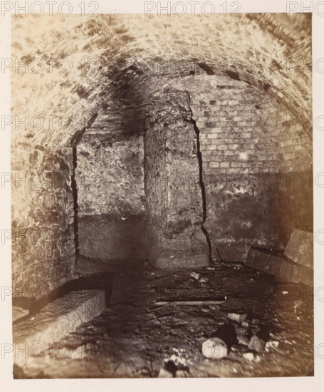 Interior of the former printing house of Johannes Gutenberg in Mainz, photograph, page, number: '9', photographer: B. Erdmann, Mainz, c. 1867 - in or before 1872, Rhineland-Palatinate, paper, albumen print, albumendruk, height 217 mm x width 181 mm, printing, printing office, A dimly lit underground space with a low arched ceiling made of rough bricks and mortar, a central vertical column of stone showing a large irregular crack, uneven stone walls on either side with visible mortar lines, a rough stone floor strewn with scattered debris and a few larger flat stones, and areas of light and shadow emphasizing the textured, aged surfaces.