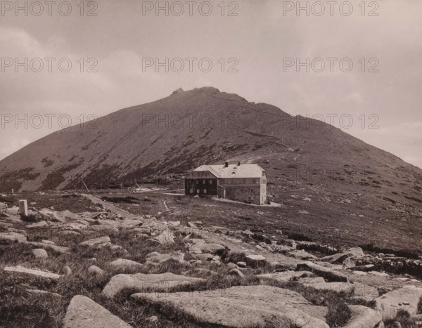 Die Schneekoppe mit der Riesenbaude, View of the Schneekoppe, page, photomechanical print, maker: E. Mertens & Cie, Giant Mountains, publisher: Max Leipelt, Warmbrunn, 1891, Silesia, Dolnoslaskie voivodship, Jelenia Góra, paper, collotype, photoprint, height 202 mm x width 268 mm, mountains, housing, façade (of house or building), Schneekoppe, A wide landscape with a rocky foreground and scattered low vegetation leading toward a gently sloping hillside, a large multi-story stone and wooden building with a pitched roof sits near the base of the hill, a narrow winding path ascends the hill toward a small structure near the summit, the hillside shows patches of bare rock and sparse low plants, the sky above is pale and mostly uniform with faint cloud formations.