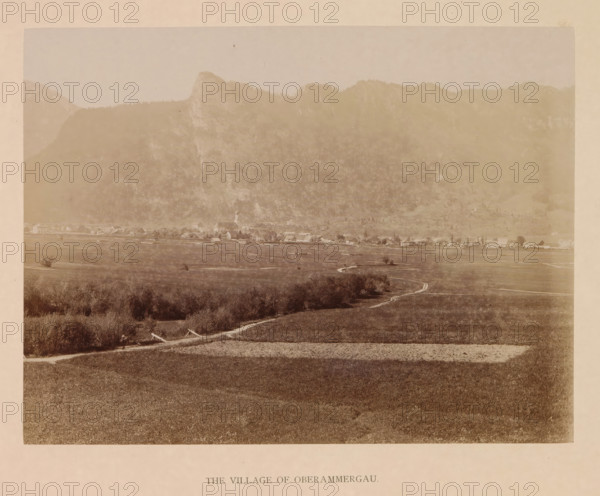 The village of Oberammergau, photograph, page, inscription: 'G. Stuffler K. Hofkunsthandlung Munich', Bavaria, c. 1885 - in or before 1890, photographer, paper, albumen print, height 168 mm x width 220 mm, city-view in general, 'veduta', Oberammergau, A wide sepia-toned landscape photograph showing a flat grassy foreground with a winding dirt path leading toward a distant village, low shrubs and small clusters of trees scattered along the field, a line of buildings with pitched roofs and a church steeple at the village edge, beyond the village a steep wooded hillside rising into craggy mountain peaks, and a pale sky above.