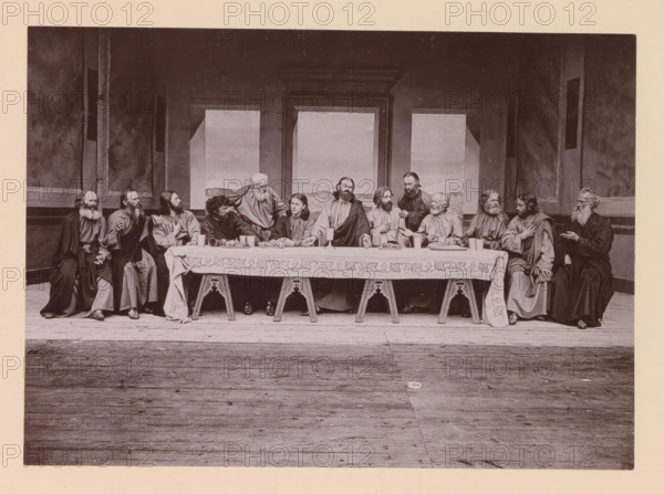 The holy supper, page, photomechanical print, inscription: 'Gemeinde Oberammergau Passionsspiel 1890', photographer: Carl Stockmann, Oberammergau, cliché maker: anonymous, c. 1885 - in or before 1890, Bavaria, paper, collotype, photoprint, height 165 mm x width 227 mm, group of actors, troupe, actors on the stage, the episode of the Last Supper, A long rectangular scene shows a group of people seated closely along one side of a long table draped with a patterned cloth, with wooden trestle legs visible beneath; the table holds plates of food, cups, and a few vessels. The people wear long robes and have varied facial hair and hairstyles, with some looking toward each other and others facing forward; one person stands behind the table leaning toward the seated figures. Behind them are tall framed wall panels with painted landscape inserts and darker surrounding walls with vertical pilasters. The floor in front of the table is wooden planks extending toward the foreground. The image is mon...
