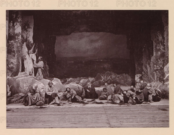 Christ's agony in the garden of Gethsemani, page, photomechanical print, inscription: 'Gemeinde Oberammergau Passionsspiel 1890', photographer: Carl Stockmann, Oberammergau, cliché maker: anonymous, c. 1885 - in or before 1890, Carl Stockmann, photographer, cliché maker, Bavaria, paper, collotype, photoprint, height 167 mm x width 227 mm, group of actors, troupe, actors on the stage, agony of Christ: to comfort him one or more angels appear to Christ with chalice and, or cross, A wide scene showing a group of robed men gathered among large rocks and plants in front of a dark cave opening, with some men seated, some reclining, and some leaning on stones; an angelic figure with wings stands on a rock near the cave entrance raising one hand while a bearded man kneels before the angel; surrounding the central figures are rocky formations, foliage, and a wooden floor in the foreground.