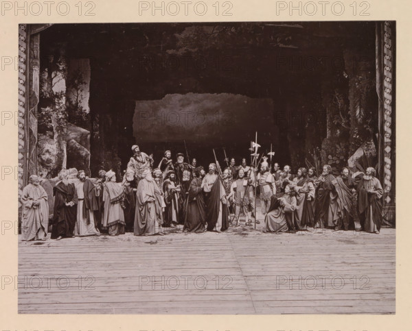 The betrayal, page, photomechanical print, inscription: 'Gemeinde Oberammergau Passionsspiel 1890', photographer: Carl Stockmann, Oberammergau, cliché maker: anonymous, c. 1885 - in or before 1890, Bavaria, paper, collotype, photoprint, height 162 mm x width 208 mm, group of actors, troupe, actors on the stage, the kiss of Judas: accompanied by soldiers with torches and lanterns, he kisses Christ, A wide stage scene framed by patterned vertical borders shows a group of people dressed in long robes and period garments gathered across the middle ground, some standing and some seated, with a few holding long pole weapons and banners; the background is dark with painted foliage and rock formations at the sides, and the foreground is a wooden floor with visible planks extending toward the viewer.