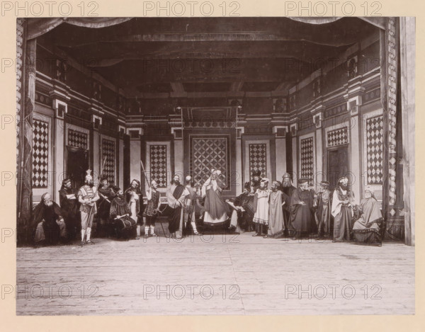 Christ before the highpriest Caiaphas, page, photomechanical print, inscription: 'Gemeinde Oberammergau Passionsspiel 1890', photographer: Carl Stockmann, Oberammergau, cliché maker: anonymous, c. 1885 - in or before 1890, Bavaria, paper, collotype, photoprint, height 162 mm x width 216 mm, group of actors, troupe, actors on the stage, Christ before the Sanhedrin with Caiaphas as high priest, and possibly Annas, maybe a soldier about to strike Christ because he keeps silent (Matthew 26:57-66, Mark 14:53-64, Luke 22:54, 22:66-71), A wide stage scene shows a group of people in historical costumes standing and sitting across an ornate set with patterned wall panels and decorative columns, a draped proscenium at the top, and a wooden floor in the foreground; the figures wear robes, tunics, helmets, cloaks, and head coverings, and some hold staffs, spears, or other props while others are seated or gesturing, with detailed geometric designs visible on the backdrop and side panels.