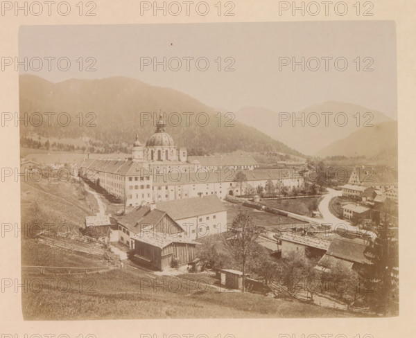 The village of Ettal, photograph, page, inscription: 'G. Stuffler K. Hofkunsthandlung Munich', Bavaria, c. 1885 - in or before 1890, photographer, paper, albumen print, height 167 mm x width 212 mm, city-view in general, 'veduta', Ettal, A sepia-toned panoramic photograph shows a valley town with a large domed church and adjacent buildings occupying the central area, surrounded by rolling hills and distant mountains shrouded in haze. The foreground contains sloping grassy fields with wooden fences, small sheds, and clusterings of rooftops and barns. A winding road curves through the scene past rows of multi-story rectangular buildings with pitched roofs and numerous windows, small trees dot the grounds, and a river or canal runs alongside part of the built area. The church features a prominent rounded dome topped by a smaller cupola and flanked by a slender tower, and terraced buildings extend from it in linear blocks. The overall image has a faded, aged appearance with soft contrast a...