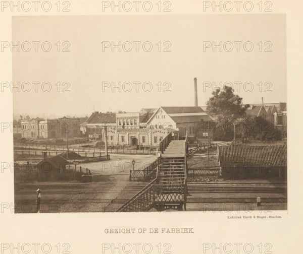 View of the factory, View of the machine factory of Stork & Co. Brothers, page, photomechanical print, number: 'No. 1.', Hengelo (Overijssel), maker: Emrik & Binger, Haarlem, c. 1884 - in or before 1894, photographer, paper, collotype, photogravure, height 154 mm x width 214 mm, Print opposite title page., factory-building, A sepia-toned photograph of an industrial waterfront scene showing a wooden pedestrian bridge with stairs and lattice railings crossing over railroad tracks toward a group of brick and masonry factory buildings, roofs of varying pitches and textures, and a tall cylindrical smokestack rising behind them; a fenced yard and small outbuildings are visible nearby, along with a large tree beside one of the factory structures, a few lamp posts and gate posts near the tracks, and distant low buildings and rooftops under a pale sky.