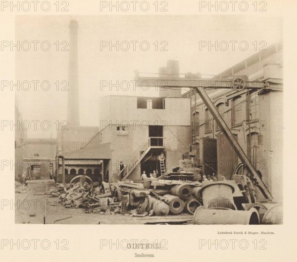 Foundry. Smelting furnaces, View of the smelting furnaces of the foundry of the machine factory of Gebroeders Stork & Co, page, photomechanical print, number: 'No. 6.', Hengelo (Overijssel), maker: Emrik & Binger, Haarlem, c. 1884 - in or before 1894, photographer, paper, collotype, photogravure, height 155 mm x width 200 mm, factory-building, fire, oven, furnace, interior of industrial structure, workshop, A sepia-toned industrial yard with piles of large metal cylinders, wheels, and assorted castings scattered across the ground in front of brick buildings; an elevated metal beam structure with a pulley wheel and supporting diagonal brace spans between buildings; a multi-story brick and corrugated structure with external stairs, ladders, and a doorway shows small groups of people standing near the entrance and on the stairs; tall chimneys and rooftop elements rise behind the buildings, and tracks and pathways run through the yard among equipment and debris.