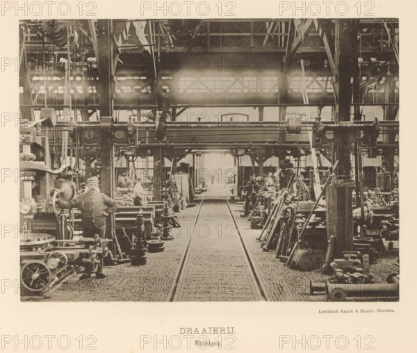 Interior with employees of the machine factory of Gebroeders Stork & Co., Turning Shop. Central aisle, page, photomechanical print, number: 'No. 9.', Hengelo (Overijssel), maker: Emrik & Binger, Haarlem, c. 1884 - in or before 1894, photographer, paper, collotype, photogravure, height 157 mm x width 210 mm, factory-building, factory-interior, machine shop, A wide interior view of an industrial workshop with a cobblestone central aisle flanked by metal tracks leading toward a distant open doorway, heavy overhead steel beams and a gallery with pulleys and belts near the ceiling, rows of large machines and gear assemblies lining both sides of the aisle, stacks of metal parts and tools arranged beside the machines, several men in work clothes standing and sitting at the machinery, a man in the foreground with one hand resting on a large machine wheel, ladders and structural columns interspersed among the equipment, and light coming through high windows near the back of the space.