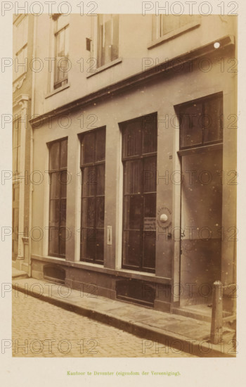 View of the office of the Geldersche Crediet-Vereeniging in Deventer, Office in Deventer (property of the Association), photograph, page, Deventer, c. 1891 - c. 1892, photographer, paper, albumen print, height 193 mm x width 126 mm, façade (of house or building), A sepia-toned street scene showing the facade of a narrow building with tall, divided windows set in a smooth plastered wall, a recessed doorway with a round door handle plate, a column-like wall edge at the corner, a raised curb and cobblestone pavement in front, narrow rectangular ventilation grilles at the base of the facade, a small metal post near the entrance, upper floor windows with sills, and subtle speckled aging marks across the photograph.
