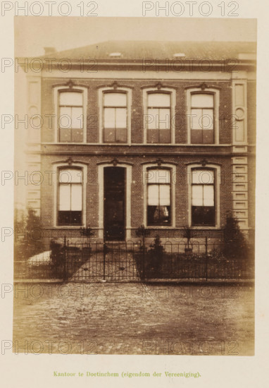 View of the office of the Geldersche Crediet-Vereeniging in Doetinchem, Office in Doetinchem (property of the Association), photograph, page, Doetinchem, c. 1891 - c. 1892, photographer, paper, albumen print, height 155 mm x width 111 mm, façade (of house or building), A two-story brick building facade with a central doorway flanked by tall rectangular windows on each side and matching windows above, decorative stone trim around the windows and along the building corners, a low iron fence and a paved path leading to the entrance, small plants and shrubs in front of the building, a tiled roof with roof windows partially visible, and a cobblestone street in the foreground.