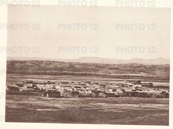 Pueblo of Santo Domingo, page, photomechanical print, number: 'Fig. 22.', photographer: John Karl Hillers, New Mexico, cliché maker: Heliotype Printing Company, Boston (Massachusetts), c. 1876 - in or before 1881, North America, Massachusetts, paper, collotype, photoprint, height 160 mm x width 221 mm, city-view in general, 'veduta', Santo Domingo Pueblo, A wide, sepia-toned landscape showing a flat foreground with textured ground leading to a clustered settlement of low, rectangular buildings with flat roofs, some taller structures rising slightly above the rest; behind the settlement there is a narrow strip of lighter land and rows of vegetation, then rolling hills with mottled, bush-covered slopes, and a distant line of low mountains beneath a pale, empty sky.