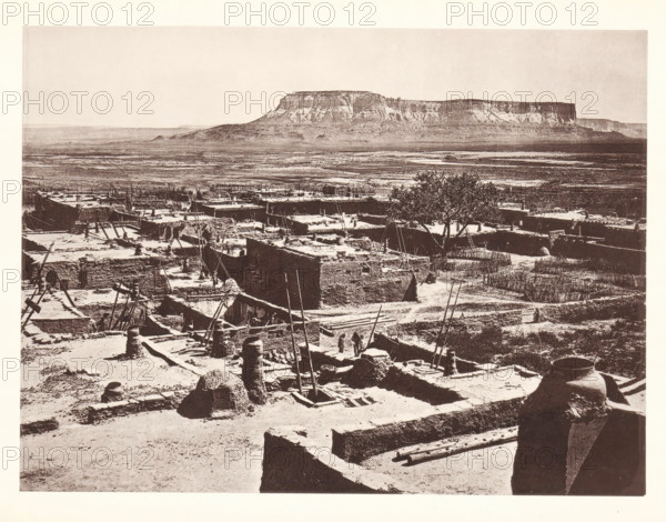 Pueblo of Zuni, page, photomechanical print, number: 'Fig. 23.', photographer: John Karl Hillers, New Mexico, cliché maker: Heliotype Printing Company, Boston (Massachusetts), c. 1876 - in or before 1881, North America, Massachusetts, paper, collotype, photoprint, height 172 mm x width 231 mm, city-view in general, 'veduta', Zuni Pueblo, A wide sepia-toned scene shows a sprawling adobe village with flat-roofed mud structures, many with scaffolding poles and ladders leaning against them, and narrow pathways winding between the buildings; in the foreground a large rounded clay jar sits on a ledge beside stacked rectangular walls and scattered wooden beams, while further back a taller tree rises above the rooftops and a distant flat-topped mesa dominates the horizon over an open plain.
