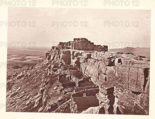 Pueblo of Wolpi, page, photomechanical print, number: 'Fig. 25.', photographer: John Karl Hillers, Arizona, cliché maker: Heliotype Printing Company, Boston (Massachusetts), c. 1876 - in or before 1881, North America, Massachusetts, paper, collotype, photoprint, height 160 mm x width 216 mm, city-view in general, 'veduta', mountains, Walpi, A sepia-toned photograph shows a rocky mesa with layered cliff faces and rough stone terraces leading up to a cluster of rectangular stone structures perched at the top; the foreground features steep, eroded rock with small recessed openings and stacked stone enclosures, while the midground reveals the fortified buildings and the background opens to a broad, flat plain under a pale sky.