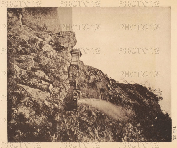 View of a steam train on the track between La Guaira and Caracas in Venezuela, page, photomechanical print, number: 'Tab. XX.', photographer: Jacobus Rudolphus Neervoort van de Poll (possibly), Venezuela, cliché maker: anonymous, c. 1877 - in or before 1887, paper, collotype, photoprint, height 165 mm x width 136 mm, Print in section at the back., railway, train, A sepia-toned photograph showing a rocky cliffside with layered textured rock surfaces and sparse vegetation. A narrow wooden walkway or bridge with railings is attached along the face of the cliff, leading toward a small platform or structure perched near a rock outcrop. Wisps of mist or spray rise along the cliff near the walkway. The sky appears empty and pale above the cliff, and the image has a worn, antique appearance with a narrow border around it.
