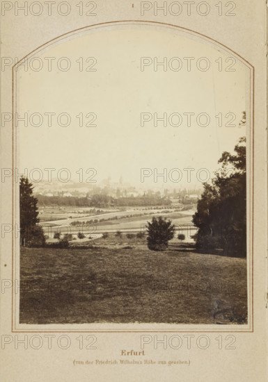 View of Erfurt, Erfurt (von der Friedrich Wilhelm's Höhe aus gesehen), photograph, page, number: '1', Erfurt (district), c. 1858 - in or before 1868, photographer, Thuringia, paper, albumen print, height 128 mm x width 98 mm, Image on frontispiece., city-view in general, 'veduta', Erfurt (city), A sepia-toned framed photograph showing a wide landscape from a slightly elevated viewpoint, with a foreground of grassy terrain that slopes gently toward a middle ground of open fields and scattered small trees and shrubs, a solitary bush near center foreground, and a winding lighter-toned path or road traversing the fields; taller trees flank the scene on the left and right edges, partially obscuring the view, while the distant background fades into a hazy skyline with faint outlines of clustered buildings and distant church spires barely visible against a pale sky.