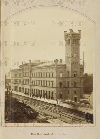 View of the Erfurt train station, Das Bahnhof-Gebäude, photograph, page, number: '8.', Erfurt (city), c. 1858 - in or before 1868, photographer, Thuringia, paper, albumen print, height 128 mm x width 98 mm, Image opposite page 100., station, shelter (railway, tramway), facade (of house or building), A sepia-toned photograph of a large stone building with rows of rectangular windows and decorative cornices, featuring a tall square clock tower with a crenellated top and round clock faces, a street-level platform beside multiple sets of railroad tracks with several open rail cars visible, small groups of people standing and walking along the platform, scaffolding attached to part of the tower, and trees visible in the background.