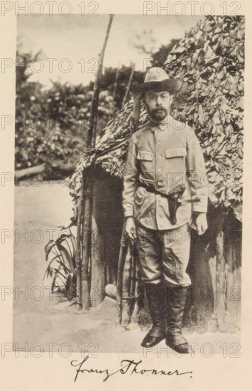 Portrait of Franz Thonner in front of a wooden house, Franz Thonner, page, photomechanical print, cliché maker: Stengel & Markert, Dresden, 1896, photographer, Saxony, paper, collotype, photogravure, height 162 mm x width 109 mm, Print after page X., Person, explorer, A sepia-toned photograph of a bearded man standing in front of a thatched structure with leafy covering, wearing a wide-brimmed hat, a buttoned jacket with flap pockets, a belt at the waist, trousers tucked into tall boots, and holding a long wooden staff; behind him are vertical poles and dense foliage, and a patch of ground and plants is visible at his feet.