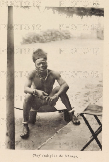 Chef indigène de Mbinga, Portrait of the chieftain of Mbinga, page, photomechanical print, number: 'Pl. 80.', photographer: Franz Thonner, Mbinga, cliché maker: Stengel & Markert, Dresden, 1896, Saxony, paper, collotype, photoprint, height 161 mm x width 110 mm, ruler, sovereign, A sepia-toned photograph showing a shirtless man seated on a low wooden stool under a thatched roof edge visible at the top; he wears a tall fur cap, beaded necklaces, bracelets, and anklets, and holds a rounded object in his hands while looking toward the camera; a vertical post stands to the left of him, the ground beneath him is sandy, a blurred cluster of foliage appears in the background, and part of a wooden structure is visible at the lower right.