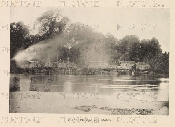 Etebe, village des Mobali, View of Etebe, page, photomechanical print, number: 'Pl. 45.', photographer: Franz Thonner, Congo (country), cliché maker: Stengel & Markert, Dresden, 1896, Saxony, paper, collotype, photoprint, height 110 mm x width 161 mm, city-view in general, 'veduta', façade (of house or building), A riverside scene shows a row of thatched huts and raised structures along the water's edge, with conical and rounded roofs made of straw or similar material and wooden stilts visible beneath some huts; dense trees and foliage rise behind the huts, forming a dark treeline with taller tree canopies toward the left; the river in front reflects the huts and trees with gentle ripples, and remnants of wooden posts or stumps project from the shallow water nearer the shore; a sandy foreground contains scattered bundles of reeds or straw and a shallow bowl or basin resting on the sand; a soft mist or spray drifts across the left side of the image, blurring part of the huts and trees.