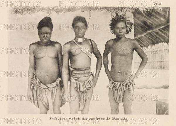 Portrait of three Mobali men near Monveda, Indigènes mobali des environs de Monveda, page, photomechanical print, number: 'Pl. 46.', photographer: Franz Thonner, Congo (country), cliché maker: Stengel & Markert, Dresden, 1896, Saxony, paper, collotype, photoprint, height 108 mm x width 162 mm, adult man, Three dark-skinned individuals stand side by side beneath a thatched roof, each wearing minimal cloth skirts and bare chests; the person on the left has a taller hair arrangement and a rounded abdomen, the central figure wears a strap across the chest and a small rectangular ornament at the neck, and the person on the right has short hair adorned with foliage and a hand resting on the hip, while a bamboo or wooden fence and a sloped thatched wall are visible in the background.