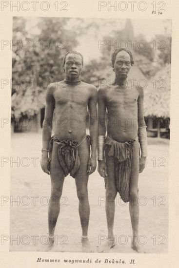 Hommes mogwandi de Bokula II, Portrait of two men from the Ngbandi at Bokula, page, photomechanical print, number: 'Pl. 74.', photographer: Franz Thonner, Bokula, cliché maker: Stengel & Markert, Dresden, 1896, Saxony, paper, collotype, photoprint, height 163 mm x width 110 mm, adult man, A sepia-toned photograph of two shirtless men standing side by side facing the camera, both wearing simple loincloths and ankle bands, with one man wearing multiple stacked metal bracelets on both forearms and the other wearing a single bracelet on one wrist; both men have short hair with visible decorative markings or scars on their foreheads and chests, and they stand on bare ground with blurred vegetation and a thatched hut visible in the background.