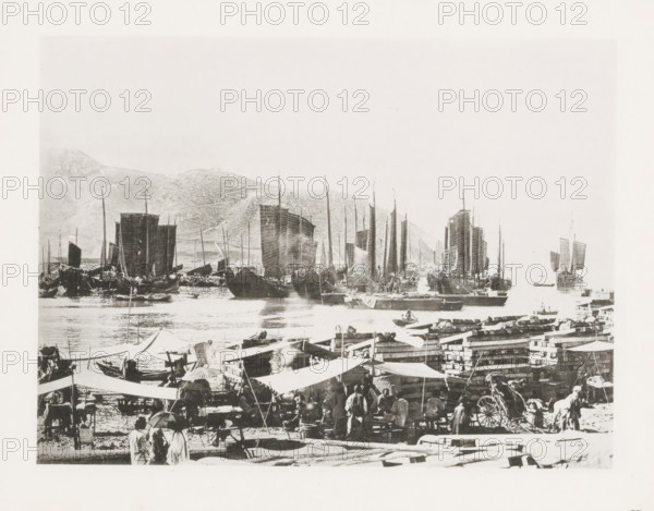 Drying place for sails in Port Arthur, with Chinese junks in the background, page, photomechanical print, number: '15', photographer: AN Ljupova, Port Arthur, cliché maker: anonymous, 1898 - 1899, Manchuria, paper, collotype, photoprint, height 150 mm x width 203 mm, ships (in general), sail, A busy waterfront scene with numerous traditional sailing vessels lined up on calm water, their tall masts and large rectangular sails prominent against a pale sky and distant hilly shoreline; closer to the viewer a crowded quay area shows market stalls and canvas awnings, stacks of crates and timber, and groups of people standing and moving about, some wearing wide-brimmed hats and carrying umbrellas, while small boats and rowboats dot the water near the larger ships.