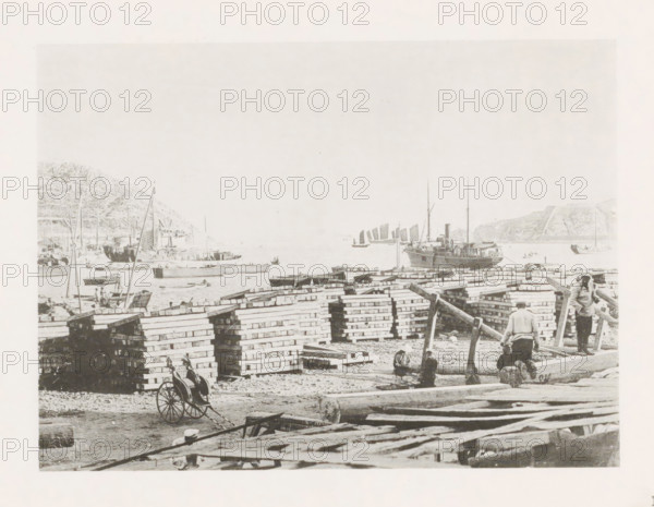 Quay in Port Arthur, with a view of the bay, page, photomechanical print, number: '16', photographer: AN Ljupova, Port Arthur, cliché maker: anonymous, 1898 - 1899, Manchuria, paper, collotype, photoprint, height 150 mm x width 203 mm, gulf, bay, pier, quay, wharf, rickshaw, A waterfront scene with stacks of rectangular wooden crates arranged in rows across the shore, long logs and planks scattered in the foreground, and a handcart with large spoked wheels near the crates; small groups of people are standing, sitting, and working among the lumber, some on a large log and others near the waterline; several small boats and larger sailing vessels with tall masts are moored or afloat in the bay, and a steamship is anchored closer to the right; hills rise on both sides of the bay in the background.