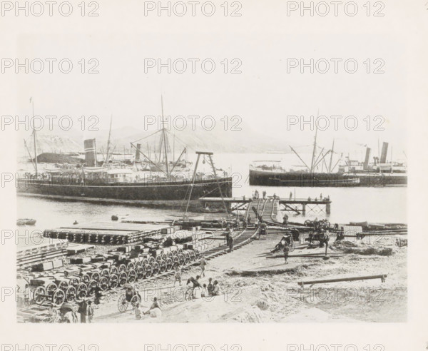 Jetty of the East China Railway at Port Arthur, page, photomechanical print, number: '17', photographer: AN Ljupova, Port Arthur, cliché maker: anonymous, 1898 - 1899, Manchuria, paper, collotype, photoprint, height 150 mm x width 203 mm, railway, tramway, rack railway, ships (in general), A harbor scene showing several large steamships moored in the water with tall masts and funnels, a wooden pier extending out with people standing and walking on it, a crane-like structure near the pier, rows of wagon wheels and stacked crates or beams arranged on the shore, horse-drawn carts and scattered figures of people working or resting on the beach, a track or wooden rails leading from the shore onto the pier, and distant low hills visible across the water.