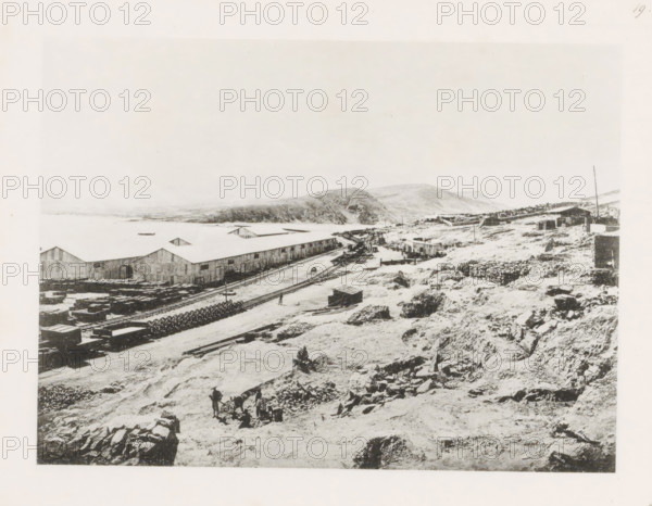 View of the East China Railway, page, photomechanical print, number: '19', photographer: AN Ljupova, Manchuria, cliché maker: anonymous, 1898 - 1899, paper, collotype, photoprint, height 150 mm x width 203 mm, warehouse (, harbour), railway, tramway, rack railway, A wide black-and-white scene showing a rocky, uneven shoreline with scattered stones and rough ground in the foreground, a few small groups of people working among tools and carts, a set of parallel railway tracks running along the midground beside long warehouse buildings with peaked roofs, stacks of materials and circular coils near the tracks, a curved dirt road leading toward a cluster of low buildings and structures further back, rolling hills rising behind the buildings, and a pale sky above.