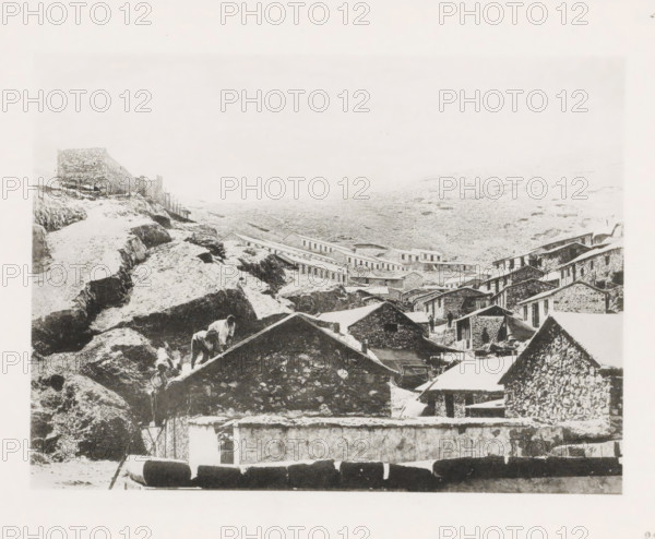 Workers' houses along the slope of a mountain in Port Arthur, page, photomechanical print, number: '23', photographer: AN Ljupova, Port Arthur, cliché maker: anonymous, 1898 - 1899, AN Ljupova, photographer, cliché maker, Manchuria, paper, collotype, light print, height 150 mm x width 203 mm, working class, labourers, (high) hill, housing, facade (of house or building), A sepia-toned scene shows a cluster of stone buildings with sloped roofs arranged on a rocky hillside, with terraces and retaining walls visible among rough outcrops. A steep rocky path winds up the left side toward a stone wall with a small structure, and a row of low long buildings stretches across the midground along the slope. In the foreground several pitched roofs and stone facades form a dense arrangement, with a few indistinct human figures near the buildings and someone on a roof. The landscape behind the settlement is a barren, gently sloping expanse with scattered rocks, and the sky above is pale and largely ...