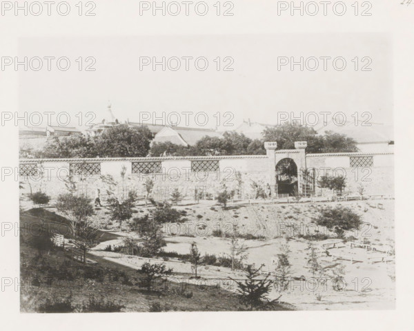 View of the Nicholas Boulevard in Port Arthur, page, photomechanical print, number: '24', photographer: AN Ljupova, Port Arthur, cliché maker: anonymous, 1898 - 1899, Manchuria, paper, collotype, light pressure, height 150 mm x width 203 mm, city-walls, avenue, boulevard in village, A wide view shows a long stone wall with decorative lattice sections and a central arched gateway topped by short battlement-like structures, behind which tree canopies rise and tiled roofs with ornate ridges are visible; in front of the wall there is an embankment with scattered small trees and shrubs, simple wooden fences and walkways, and a few people near the gateway, all rendered in a faded monochrome tone.