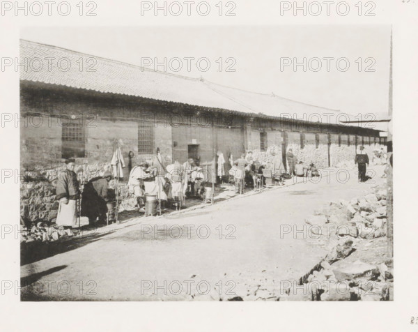 Street barbers in Port Arthur, page, photomechanical print, number: '25', photographer: AN Ljupova, Port Arthur, cliché maker: anonymous, 1898 - 1899, Manchuria, paper, collotype, photoprint, height 150 mm x width 203 mm, street, barber, barbershop, shaving, being shaved, A long single-story building with a tiled roof and stone lower walls extends along a street, with several rectangular windows and a central doorway; a line of people stand and sit along the building’s side, some wearing cloaks and hats and others with aprons, gathered around buckets, stools, and small stands; clothing and bundled items hang on hooks or lean against the wall; a man walks along the street away from the group toward the right side of the image; piles of stones and rubble are scattered along the curb in the foreground and near the right edge; shadows fall across the street and the overall scene appears to be in monochrome.