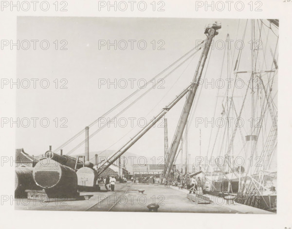 Port crane at Port Arthur, page, photomechanical print, number: '26', photographer: AN Ljupova, Port Arthur, cliché maker: anonymous, 1899 - 1898, Manchuria, paper, collotype, photoprint, height 150 mm x width 203 mm, harbour, dock crane, A dock scene showing large cylindrical metal objects with perforated ends stacked on the left, a long angled crane boom extending upward from the dock toward the right, multiple rigging lines and masts of ships rising along the right edge, workers and figures standing and walking along the pier, tracks embedded in the pier surface, bollards and wooden planks on the dock, industrial buildings and chimneys in the background, and distant hills beneath a pale sky.