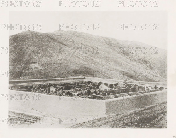 Burned Chinese camp near Port Arthur, page, photomechanical print, number: '28', photographer: AN Ljupova, Manchuria, cliché maker: anonymous, 1898 - 1899, paper, collotype, photoprint, height 150 mm x width 203 mm, ruin of a building, architecture, mountains, A grainy black-and-white photograph showing a low stone wall or rectangular enclosure in the foreground with rough, irregular stones and sections of smoother masonry, and behind it a large sloping hillside covered in textured, mottled terrain extending across the image under a pale sky.