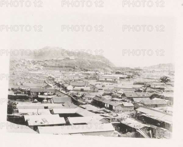 View of Port Arthur, page, photomechanical print, number: '6', photographer: AN Ljupova, Port Arthur, cliché maker: anonymous, 1898 - 1899, Manchuria, paper, collotype, photoprint, height 150 mm x width 203 mm, city-view in general, 'veduta', facade (of house or building), A wide grayscale view shows a densely packed town with many low buildings and tiled roofs extending toward a shoreline, with open sandy areas and scattered small boats near the water; a hillside rises behind the town with terraced slopes and a few isolated structures, and distant cliffs frame the right edge of the scene, while narrow streets and alleys run between rows of houses with occasional groups of small figures visible among them.