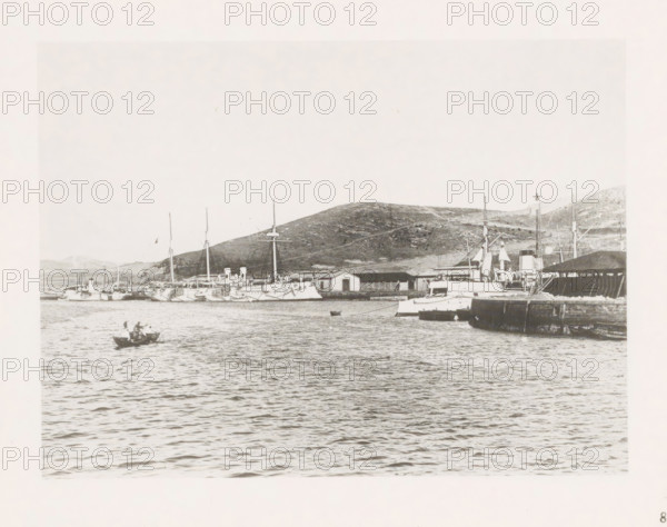 View of boats on the quay in Port Arthur, page, photomechanical print, number: '8', photographer: AN Ljupova, Port Arthur, cliché maker: anonymous, 1898 - 1899, Manchuria, paper, collotype, photoprint, height 150 mm x width 203 mm, ships (in general), pier, quay, wharf, sea, A waterfront scene with multiple moored ships and boats along a quay backed by low hills, a small rowboat with two people in the foreground on rippling water, larger vessels with masts and smokestacks lined near warehouses and a pier, and rocky terrain rising behind the harbor.
