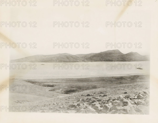 View of the bay at Port Arthur, page, photomechanical print, number: '9', photographer: AN Ljupova, Manchuria, cliché maker: anonymous, 1898, paper, collotype, photoprint, height 150 mm x width 203 mm, gulf, bay, sea, A wide, pale landscape photograph showing a calm body of water bordered by distant rolling hills and small mountains across the horizon, with a shoreline in the foreground that transitions from rocky terrain to flatter, grassy ground; two small boats float on the water near the right side and another small vessel is visible farther left; the sky above is light and largely empty, with subtle discoloration marks near the upper corners and faint variations in tone across the image.