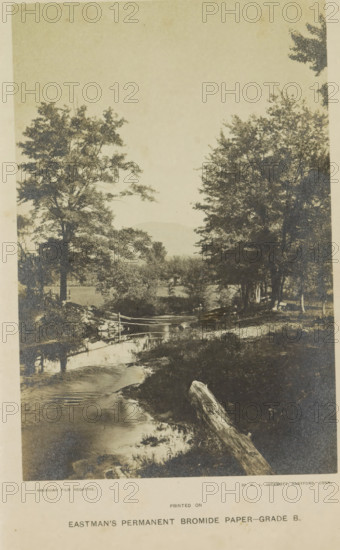 View of a ditch (presumably) in a park, photograph, page, photographer: WJ Hickmott, Connecticut, c. 1884 - c. 1889, North America, paper, gelatin silver print, height 164 mm x width 108 mm, public gardens, park, A sepia-toned landscape photograph showing a narrow stream flowing toward the foreground with reflective water and a fallen log lying along the near bank, trees with dense foliage flanking both sides of the stream, a simple wooden fence crossing the stream on a small bridge, grassy and brushy banks with scattered rocks, and distant low hills visible through the trees under a pale sky.