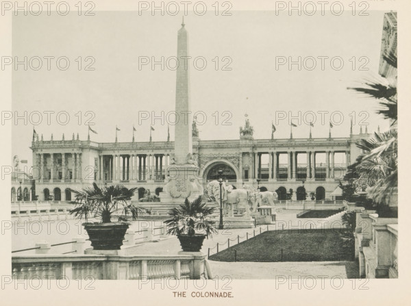 The colonnade, View of a colonnade and an obelisk at the World's Columbian Exposition in Chicago in 1893, page, photomechanical print, number: 'Plate 20', photographer: Charles Dudley Arnold, Chicago, photographer: HD Higinbotham, cliché maker: anonymous, 1893, Charles Dudley Arnold, HD Higinbotham, photographer, cliché maker, North America, Illinois, paper, collotype, photoprint, height 133 mm x width 191 mm, colonnade, obelisk, needle, world fair, world exhibition, World's Columbian Exposition, A wide plaza with a tall obelisk at its center stands in front of a large neoclassical building with a long colonnade and decorative balustrade; flagpoles line the roof and sculptural figures sit atop sections of the structure. In front of the obelisk is a raised sculptural group including a pair of large animal figures and ornate pedestals, flanked by planters containing palm-like plants. A formal path and low stone fencing run across the foreground, with trimmed grassy areas bordered by shor...