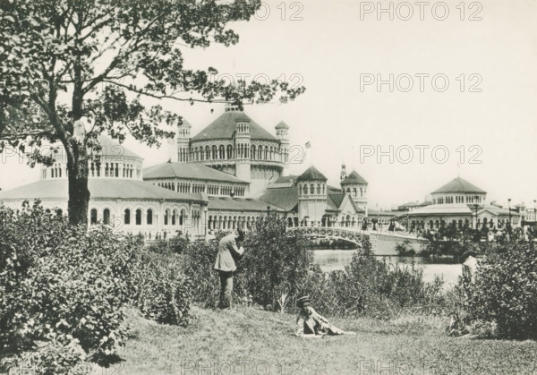 Fisheries Builing from Wooded Island, View of the fishery building, designed by Henry Ives Cobb at the World's Columbian Exposition in Chicago in 1893, page, photomechanical print, number: 'Plate 53', photographer: Charles Dudley Arnold, Chicago, photographer: HD Higinbotham, cliché maker: anonymous, 1893, Charles Dudley Arnold, HD Higinbotham, photographer, cliché maker, North America, Illinois, paper, collotype, photoprint, height 132 mm x width 190 mm, world fair, world exhibition, World's Columbian Exposition, A black and white photograph showing a large ornate building complex with domes, arched windows, and towered structures along a waterfront, partially obscured by a large tree and dense shrubbery in the foreground; a curved pedestrian bridge spans the water near the center, and people are visible on the bridge and along the shoreline; in the grassy foreground a person stands among the bushes while another person reclines on the grass wearing a hat; the sky appears light and em...