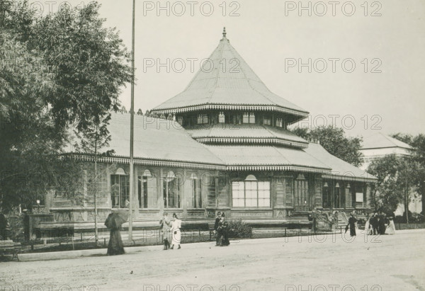 Pavilion of Ceylon at the World's Columbian Exposition in Chicago in 1893, The Ceylon Building, page, photomechanical print, number: 'Plate 79', photographer: Charles Dudley Arnold, Chicago, photographer: HD Higinbotham, cliché maker: anonymous, 1893, Charles Dudley Arnold, HD Higinbotham, photographer, cliché maker, North America, Illinois, paper, collotype, photoprint, height 134 mm x width 190 mm, world fair, world exhibition, World's Columbian Exposition, A large wooden pavilion with a wide, multi-tiered roof and decorative eaves occupies the center of the image, flanked by trees on either side and a tall slender pole in front; the building shows multiple tall windows and ornate carved details along its base and cornices, with a conical central roof topped by a finial. In front of the pavilion a few people in period clothing stand and walk along a paved area, some holding umbrellas and others gathered in small groups, while low fencing and a bench are visible near the building and ...