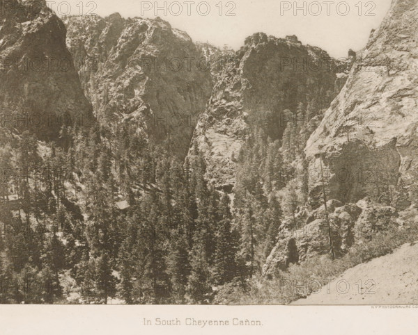 In South Cheyenne Cañon, View of the mountains near South Cheyenne Canyon, page, photomechanical print, photographer: WH Jackson Photograph and Publishing Co., Colorado Springs, cliché maker: New York Photo-Gravure Company, New York (city), c. 1888 - in or before 1893, North America, Colorado, paper, photoaquatint, height 161 mm x width 201 mm, Print opposite title page., landscapes, mountains, A sepia-toned photograph of a rugged canyon scene showing steep rocky cliffs rising from a densely forested valley, with tall evergreen trees filling the lower areas and clinging to ledges, rough textured rock faces with visible strata and crevices, a sloping foreground of lighter rocky ground at the bottom right, and a pale sky above.