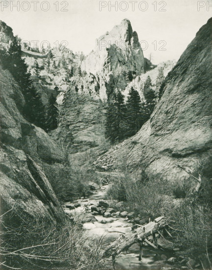 In North Cheyenne Cañon, Mountain landscape in North Cheyenne Cañon Park, page, photomechanical print, photographer: WH Jackson Photograph and Publishing Co., North Cheyenne Canyon, cliché maker: New York Photo-Gravure Company, New York (city), c. 1888 - in or before 1893, North America, Colorado, El Paso (Colorado), paper, photoaquatint, height 202 mm x width 158 mm, landscapes, mountains, A tall rocky canyon scene with steep textured rock faces on either side and a prominent jagged peak near the center; evergreen trees dot the slopes and cling to rocky ledges; a narrow stream winds through the rocky bottom with visible stones and a fallen log across the water; dry shrubs and grasses grow along the stream banks and on ledges; the sky above is pale and unobstructed.