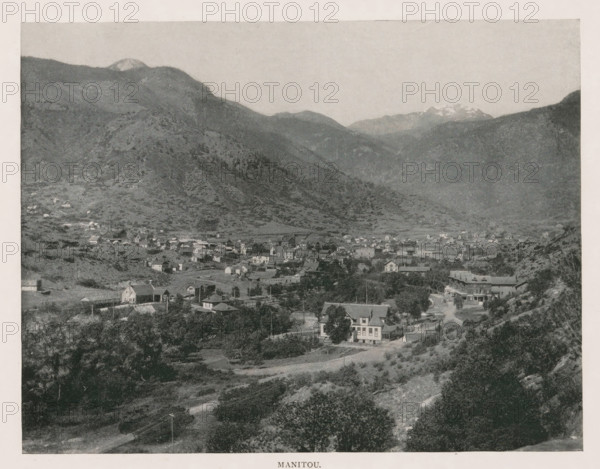 Manitou, View of Manitou Springs, page, photomechanical print, Manitou Springs, cliché maker: New York Photo-Gravure Company, New York (city), c. 1888 - in or before 1893, photographer, North America, Colorado, El Paso (Colorado), paper, autotypie, height 154 mm x width 201 mm, city-view in general, 'veduta', mountains, A wide landscape photograph shows a valley town nestled among rolling hills and steep mountains, with numerous small buildings and houses spread across the flat area and lower slopes, a few larger structures standing out, tree clusters and open grassy areas interspersed between the buildings, a road winding through the foreground past houses and vegetation, rocky hillsides and scattered shrubs rising toward the base of larger, darker mountain ridges that dominate the middle ground, and distant higher peaks visible along the horizon beneath a pale sky.