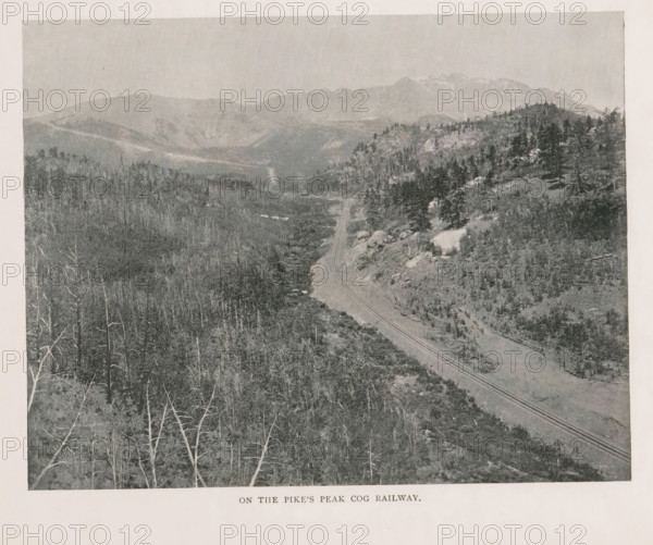 View of a cloud cover from Pikes Peak, Above the clouds, summit of Pike's Peak, page, photomechanical print, Colorado, cliché maker: New York Photo-Gravure Company, New York (city), c. 1888 - in or before 1893, New York Photo-Gravure Company, photographer, cliché maker, North America, paper, autotypie, height 90 mm x width 163 mm, clouds, mountain-top, peak, El Paso (Colorado), Pikes Peak (El Paso), A grayscale landscape photograph showing a winding railway track running through a valley with rocky slopes and scattered trees, a mix of bare trunks in the foreground and denser coniferous stands on the hillsides, with distant layered mountain ridges and a pale sky above.