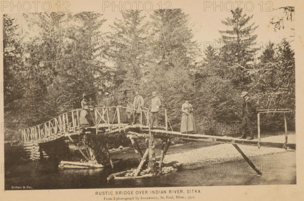 Group portrait of a group on a bridge in Sitka, Rustic bridge over Indian river, Sitka, page, photomechanical print, inscription: 'From photograph by Ingersoll, St. Paul, Minn., 3511.', photographer: Truman Ward Ingersoll, cliché maker: Britton & Rey, San Francisco, c. 1885 - in or before 1890, North America, California, Alaska, paper, collotype, photoprint, height 112 mm x width 185 mm, river, anonymous historical persons depicted in a group, in a group-portrait, A sepia-toned photograph shows a rustic wooden footbridge spanning a shallow river with visible supports and a railing made of rough logs and branches; several people stand on the bridge wearing period clothing including long dresses, coats, hats, and one person holding an umbrella, while one figure stands at the far end near a simple railing; dense evergreen trees fill the background, and the riverbank and some exposed stones are visible beneath the bridge.