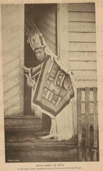 Portrait of Kitch Kawk of the Tlingit people, Kitch Kwak, of Sitka, page, photomechanical print, inscription: 'In Dancing Costume, photograph 7983, by Partridge, william henry, Portland, Oregon.', number: '7983', photographer: William Henry Partridge, Sitka, cliché maker: Britton & Rey, San Francisco, c. 1885 - in or before 1890, North America, California, Alaska, paper, collotype, photoprint, height 183 mm x width 109 mm, Person, Kitch Kawk, historical persons, city-view in general, 'veduta', glacier, A person stands in a doorway wearing a tall headdress with long upright feathers and a decorative band, a cloth draped over one shoulder, and a fringed rectangular garment with bold geometric patterns covering the front and extending toward the ground; the person has a ring in their nose and is holding the doorframe with one hand while the other hand grasps a carved object, and the scene shows wooden siding and planks around the doorway and beneath the person’s feet.